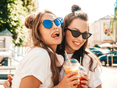 Moda kıyafetleri içinde gülümseyen iki güzel hippi kız. Dışarıda poz veren kaygısız kadınlar. Pozitif modeller, plastik bardakta pipetle taze kokteyl içiyorlar ve içiyorlar.
