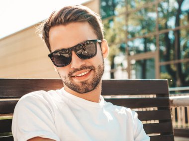 Yakışıklı, gülümseyen stil sahibi Lambersex modelinin portresi. Beyaz tişört giyen adam. Moda erkeği sokağın ortasında güneş gözlüklü bankta oturuyor.