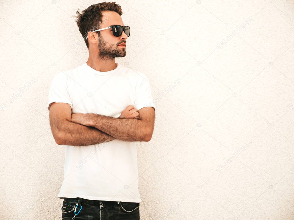 Portrait of handsome confident stylish hipster lambersexual model.Man dressed in white T-shirt. Fashion male posing on the street background near wall in sunglasses outdoors.Crossed arms