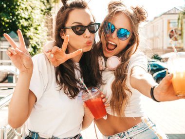 Moda kıyafetleri içinde gülümseyen iki güzel hippi kız. Dışarıda poz veren kaygısız kadınlar. Pozitif modeller, plastik bardakta pipetle taze kokteyl içiyorlar ve içiyorlar.