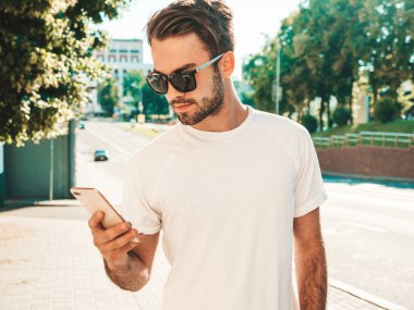 Yakışıklı, şık, havalı, lamberseksüel bir model. Beyaz tişört giymiş bir adam. Sokakta poz veren bir erkek. Cep telefonunda tutuyor, kaydırıyor ve mesaj atıyor. Akıllı telefondan mesaj yazar