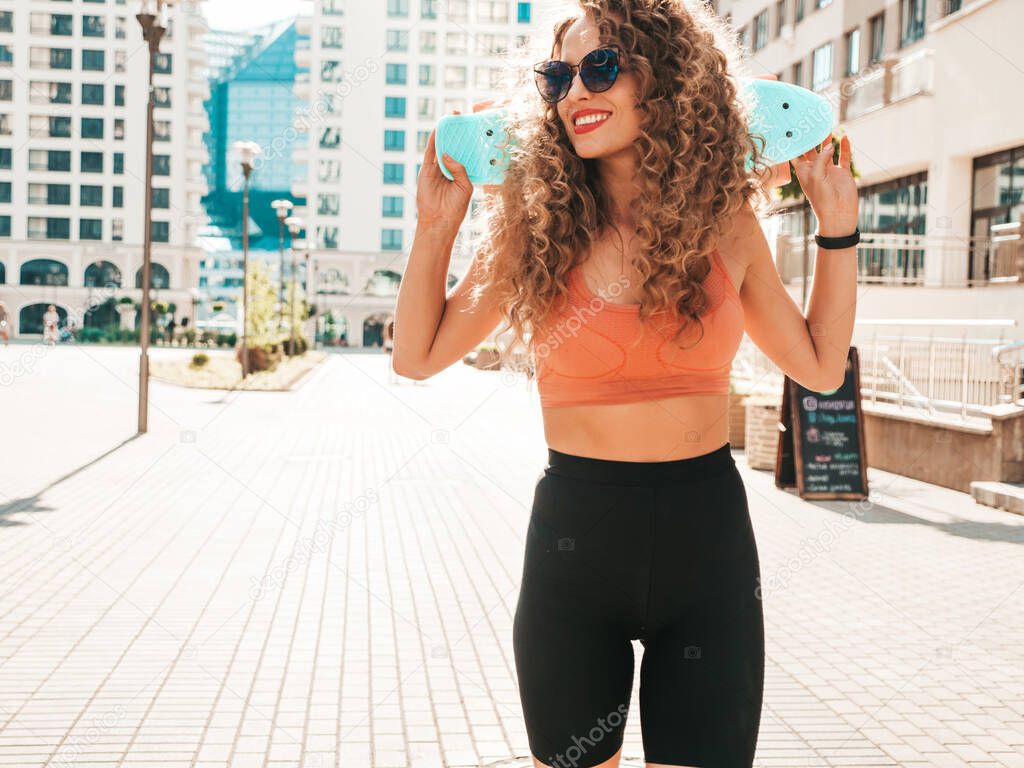 Young beautiful sexy smiling hipster woman with afro curls hairstyle.Trendy girl in summer clothes.Positive female with blue penny skateboard posing on the street background