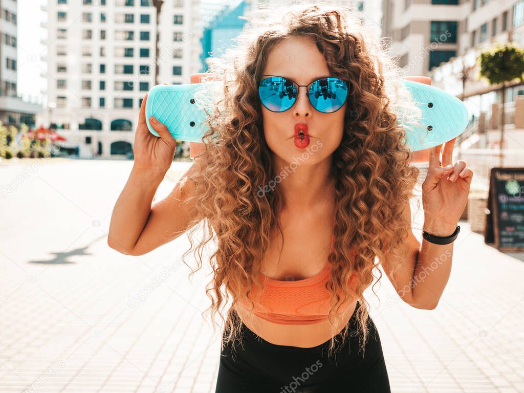 Young beautiful sexy smiling hipster woman with afro curls hairstyle.Trendy girl in summer clothes.Positive female with blue penny skateboard posing on the street background.Makes fish face