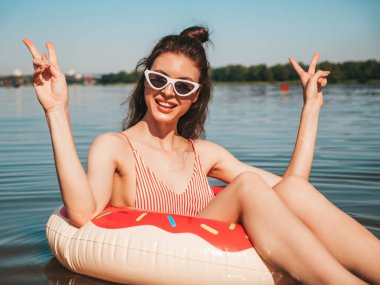 Güneş gözlüklü, genç ve seksi hippi kadın. Yazın mayo giyen, yüzen ve donut şişirilebilir yatakta yatan kadın. Göl suyunda dinlenen pozitif bir kadın. Barış işareti gösterir