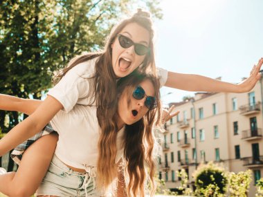 Son moda beyaz tişörtlü iki güzel gülümseyen hippi kızın portresi. Sokakta poz veren kaygısız kadınlar. Pozitif model arkadaşının sırtında oturuyor. 