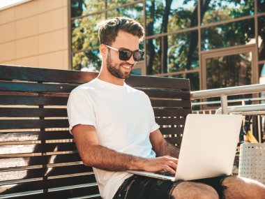 Yakışıklı, güler yüzlü, stil sahibi hipster Lambersex modeli. Moda adamı sokakta bankta oturuyor ve dizüstü bilgisayar kullanıyor, mesaj yazıyor. Ortak çalışma konsepti. Teknoloji ve iletişim