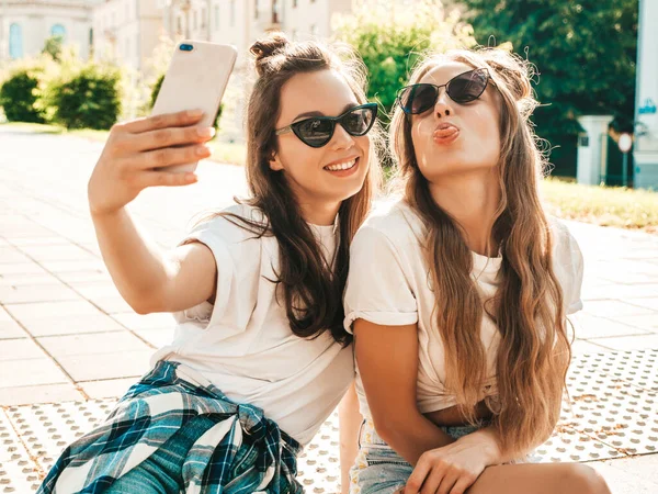 夏の服を着た2人の若い笑顔のヒップスター女性 女の子はスマートフォンで自撮り自画像写真を撮る モデルは通りに座っている 女性行くクレイジーと作る面白い顔でサングラス — ストック写真