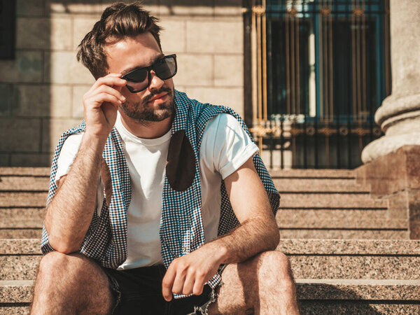 Portrait of handsome confident stylish hipster lambersexual model.Man dressed in white T-shirt. Fashion male sitting at the stairs on the street background in sunglasses