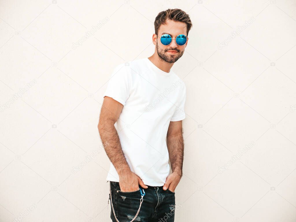 Portrait of handsome confident stylish hipster lambersexual model.Man dressed in white T-shirt. Fashion male posing on the street background near wall in sunglasses outdoors