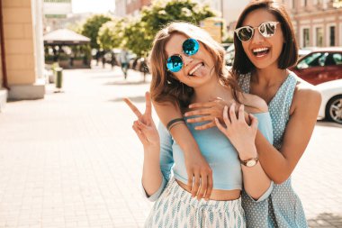 Son moda yaz kıyafetleri içinde iki güzel gülümseyen hippi kız. Güneş gözlükleriyle sokak arka planında poz veren kaygısız kadınlar. Pozitif modeller eğleniyor ve sarılıyorlar.
