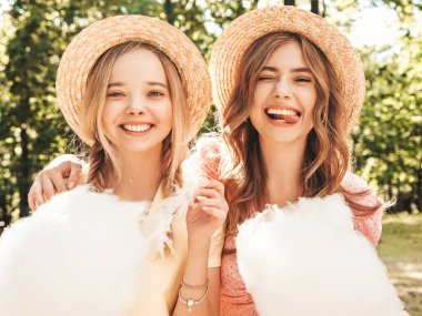 İki güzel gülümseyen yeniyetme kız şık yaz elbisesi içinde. Parkta şapkalarla poz veren kaygısız kadınlar. Pozitif modeller eğleniyor ve pamuk şeker yiyor. Kadın deliriyor