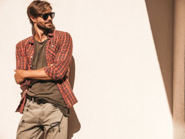 Yakışıklı, güler yüzlü, stil sahibi, lamberseksüel bir modelin portresi. Kareli gömlek giymiş bir adam. Moda erkeği sokakta güneş gözlüklü beyaz duvarın yanında poz veriyor. Kollar çapraz.