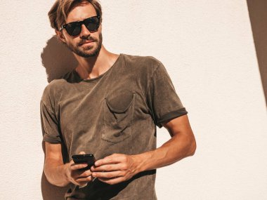 Yakışıklı, kendine güvenen, havalı, havalı, lamberseksüel model. Tişört giymiş bir adam. Beyaz duvarın yanında sokakta poz veren bir erkek. Cep telefonunda tutuyor, kaydırıyor ve mesaj atıyor. Akıllı telefondan mesaj yazar