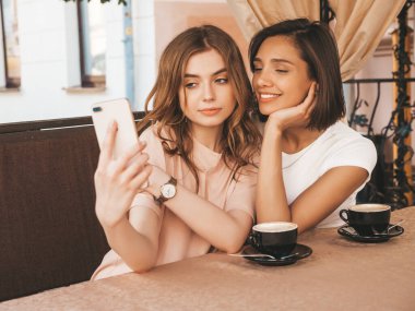 Son moda yaz kıyafetleri içinde iki güzel gülümseyen hippi kız. Kaygısız kadınlar veranda kafede sohbet edip kahve içiyor. Pozitif modeller eğleniyor ve akıllı telefondan selfie çekiyor.