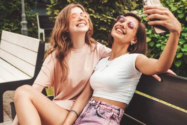 Son moda yaz kıyafetleri içinde iki güzel gülümseyen hippi kız. Sokakta güneş gözlükleriyle bankta oturan kaygısız kadınlar. Akıllı telefondan kendi portresini çekiyorlar. 