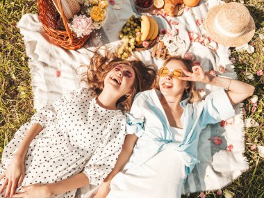 İki güzel gülümseyen yeniyetme yaz elbisesi ve şapkası içinde. Piknik yapan kaygısız kadınlar. Çimlerin üzerinde oturan, sarılan, meyve ve peynir yiyen pozitif modeller.