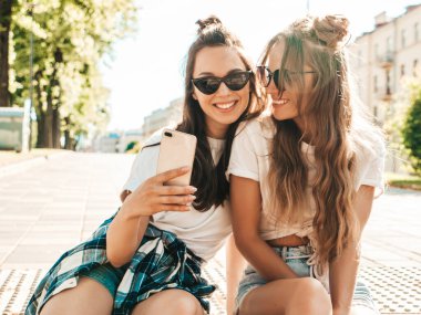 Yaz kıyafetleri içinde gülümseyen iki genç hippi kadın. Akıllı telefondan fotoğraf çeken kızlar. Sokakta oturan mankenler. Kadın çıldırıyor ve güneş gözlüklü komik suratlar yapıyor.