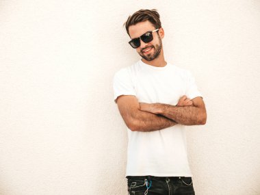 Yakışıklı, gülümseyen stil sahibi Lambersex modelinin portresi. Beyaz tişört giyen adam. Moda erkek, dışarıda güneş gözlüklü, duvarın yanında poz veriyor. Çapraz kollar