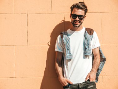 Yakışıklı, gülümseyen stil sahibi Lambersex modelinin portresi. Beyaz tişört giyen adam. Moda erkek, dışarıda güneş gözlüklü, duvarın yanında poz veriyor.