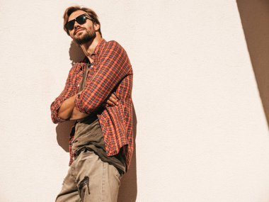 Yakışıklı, kendine güvenen, stil sahibi, Lambersex mankeni portresi. Kareli gömlek giymiş bir adam. Moda erkek, güneş gözlüklü beyaz duvarın yanında poz veriyor. Çapraz kollar