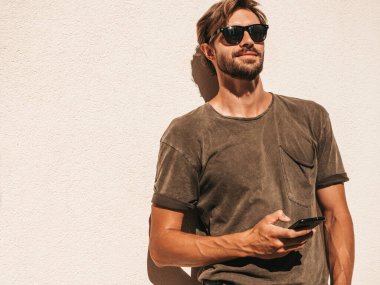 Yakışıklı, kendine güvenen, havalı, havalı, lamberseksüel model. Tişört giymiş bir adam. Beyaz duvarın yanında sokakta poz veren bir erkek. Cep telefonunda tutuyor, kaydırıyor ve mesaj atıyor. Akıllı telefondan mesaj yazıyor.