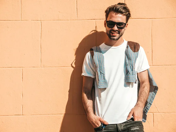 Yakışıklı, gülümseyen stil sahibi Lambersex modelinin portresi. Beyaz tişört giyen adam. Moda erkek, dışarıda güneş gözlüklü, duvarın yanında poz veriyor.