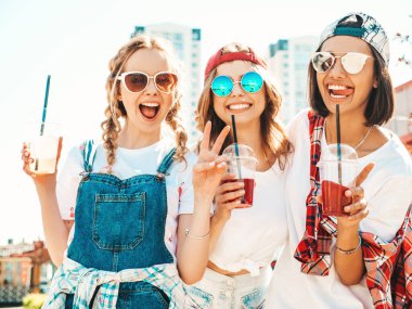 Üç güzel gülümseyen hippi kız şık yaz kıyafetleri içinde. Kaygısız kadınlar açık havada poz veriyor. Pozitif modeller, plastik bardakta pipetle taze kokteyl içiyorlar ve içiyorlar.