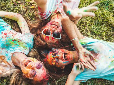 Yaz mevsiminde Holi Renk Festivali 'nde parti veren üç mutlu güzel kız. Gün batımında müzik etkinliği sonrasında gülümseyen genç bayan arkadaşlar. Güneş gözlüklü mankenler çimlerin üzerinde yatıyor.