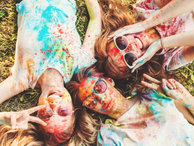 Yaz mevsiminde Holi Renk Festivali 'nde parti veren üç mutlu güzel kız. Gün batımında müzik etkinliği sonrasında gülümseyen genç bayan arkadaşlar. Güneş gözlüklü mankenler çimlerin üzerinde yatıyor.