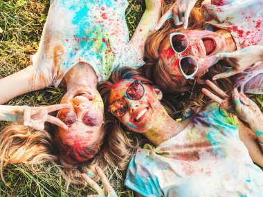 Yaz mevsiminde Holi Renk Festivali 'nde parti veren üç mutlu güzel kız. Gün batımında müzik etkinliği sonrasında gülümseyen genç bayan arkadaşlar. Güneş gözlüklü mankenler çimlerin üzerinde yatıyor.