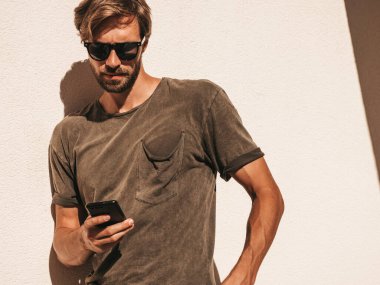 Yakışıklı, kendine güvenen, havalı, havalı, lamberseksüel model. Tişört giymiş bir adam. Beyaz duvarın yanında sokakta poz veren bir erkek. Cep telefonunda tutuyor, kaydırıyor ve mesaj atıyor. Akıllı telefondan mesaj yazıyor.