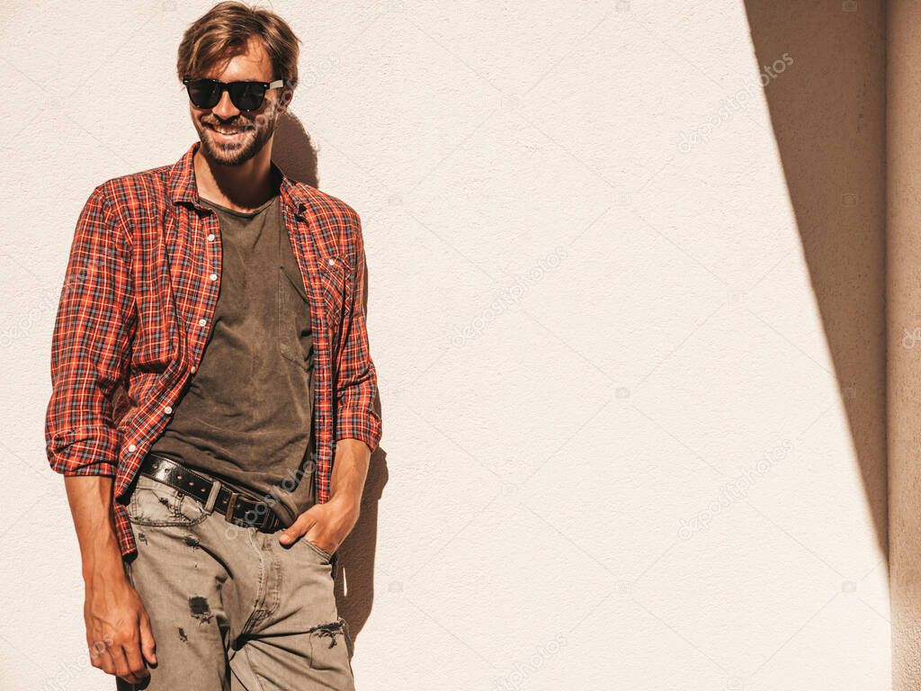 Portrait of handsome smiling stylish hipster lambersexual model.Man dressed in checkered shirt. Fashion male posing in the street near white wall outdoors in sunglasses