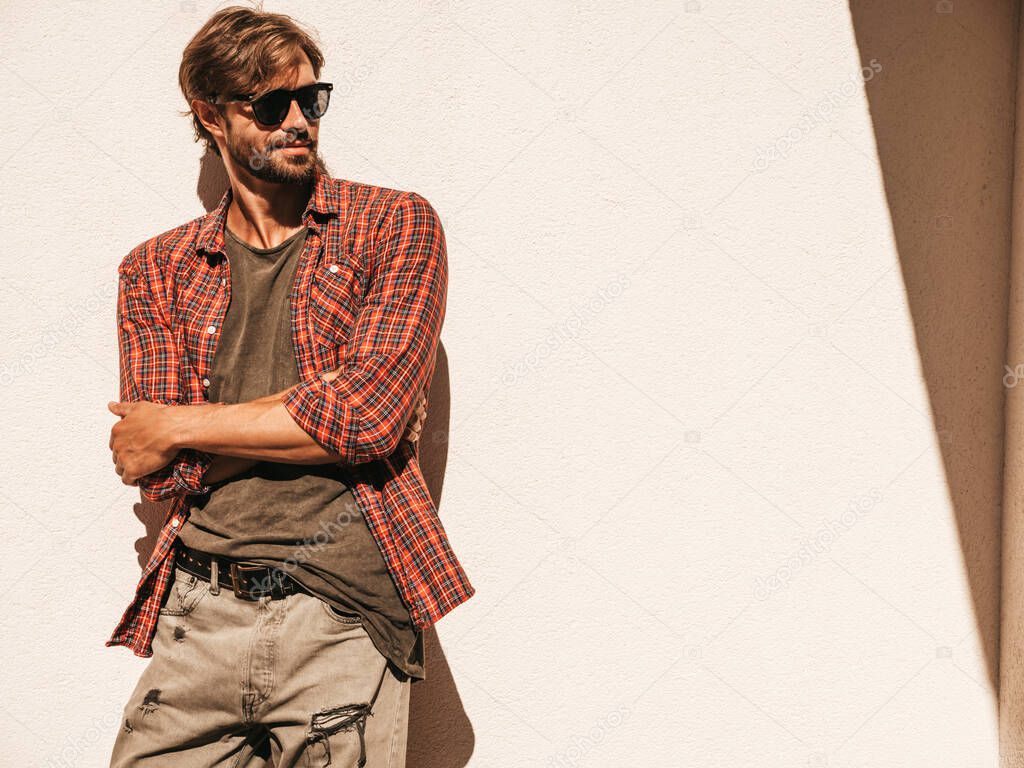 Portrait of handsome stylish hipster lambersexual model.Man dressed in checkered shirt. Fashion male posing in the street near white wall outdoors in sunglasses