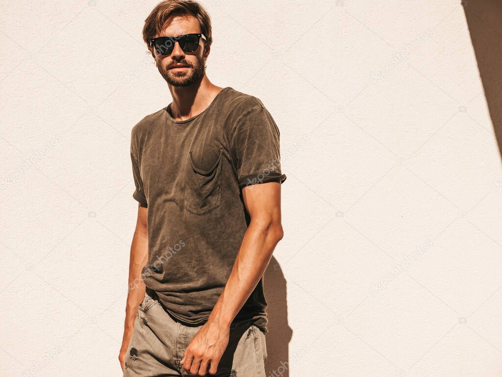 Portrait of handsome confident stylish hipster lambersexual model.Man dressed in T-shirt. Fashion male posing in the street near white wall outdoors in sunglasses