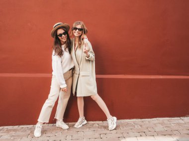 Son moda beyaz süveter ve palto giyen iki güzel, gülümseyen hippi kız. Sokakta kırmızı duvarın yanında şapkayla poz veren kaygısız seksi kadınlar. Pozitif modeller eğleniyor, güneş gözlükleriyle çıldırıyor.