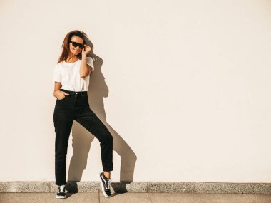 Güneş gözlüklü güzel gülümseyen modelin portresi. Hipster beyaz tişört ve kot pantolon giymiş bir kadın. Duvarın yanında poz veren modaya uygun bir kız. Komik ve pozitif kadın dışarıda eğleniyor.