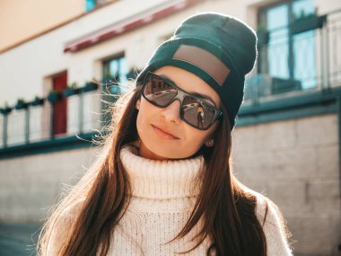 Güzel gülümseyen modelin portresi. Sıcak hippi beyaz kazak ve bere giymiş bir kadın. Günbatımında sokakta poz veren modacı kız. Komik ve pozitif kadın güneş gözlükleriyle dışarıda eğleniyor.