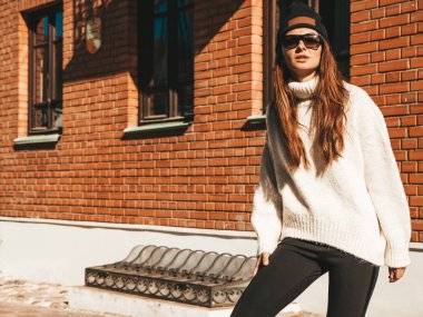 Güzel gülümseyen modelin portresi. Sıcak hippi beyaz kazak ve bere giymiş bir kadın. Tuğla binanın yanında poz veren moda bir kız. Komik ve pozitif kadın güneş gözlükleriyle eğleniyor.