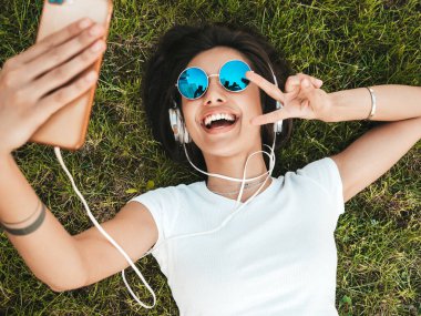 Parktaki çimlerin üzerinde yatan genç ve şık hippi kadının moda portresi. Kız modaya uygun kıyafetler giyiyor. Selfie yapıyor. Kadın kulaklıkla müzik dinliyor. Üst görünüm