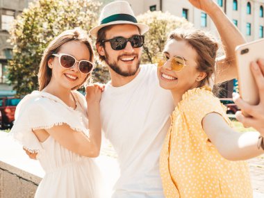 Sokakta üç şık genç arkadaş grubu. Adam ve yaz kıyafetleri giymiş iki tatlı kız. Güneş gözlükleriyle eğlenen mankenler. Kadınlar ve erkek akıllı telefondan fotoğraf çektiriyorlar.