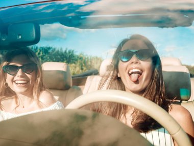Üstü açık arabadaki iki güzel ve gülümseyen hippi kızın portresi. Cabriolet kullanan kaygısız seksi kadınlar. Pozitif modeller güneş gözlükleriyle geziyor ve eğleniyorlar. Dilini gösterir