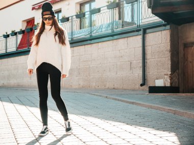 Güzel gülümseyen modelin portresi. Sıcak hippi beyaz kazak ve bere giymiş bir kadın. Günbatımında sokakta poz veren modacı kız. Komik ve pozitif kadın güneş gözlükleriyle dışarıda eğleniyor.