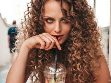 Saçları kıvırcık, güzel, gülümseyen hippi bir kadın. Yazın moda elbiseli bir kadın veranda kafede oturuyor. Model, plastik bardakta pipetle taze kokteyl içeceği içiyor.