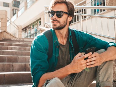 Yakışıklı, havalı, havalı, Lambersex mankeni. Beyaz tişört giymiş bir adam. Merdivenlerde oturan moda bir erkek. Cep telefonunda tutuyor, kaydırıyor ve mesaj atıyor. Akıllı telefondan mesaj yazar