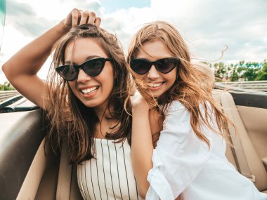Üstü açık arabadaki iki güzel ve gülümseyen hippi kızın portresi. Cabriolet kullanan kaygısız seksi kadınlar. Pozitif modeller güneş gözlükleriyle geziyor ve eğleniyorlar.