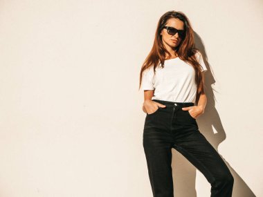 Güneş gözlüklü güzel bir modelin portresi. Hipster beyaz tişört ve kot pantolon giymiş bir kadın. Duvarın yanında poz veren modaya uygun bir kız. Komik ve pozitif kadın dışarıda eğleniyor.