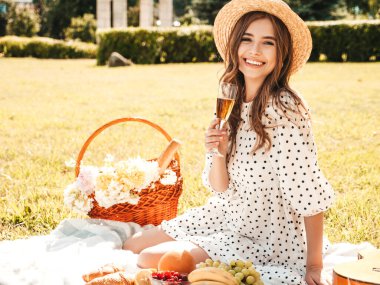 Moda elbisesi ve şapkası olan genç ve güzel hippi kadın. Kaygısız kadın dışarıda piknik yapıyor. Pozitif model çimlerin üzerinde oturuyor, meyve ve peynir yiyor, şampanya içiyor.