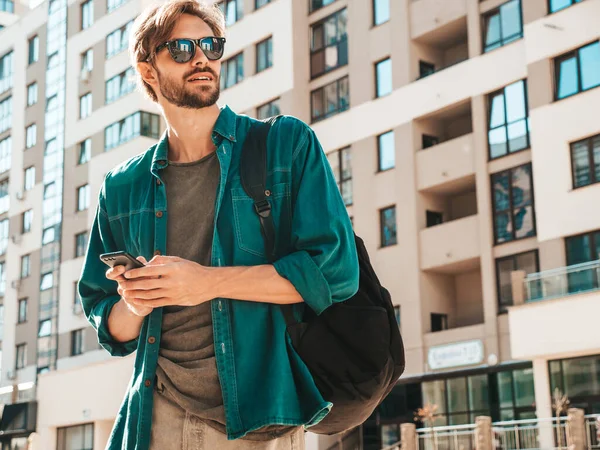 Yakışıklı, şık bir hipster Lambersex mankeni. Tişört giymiş bir adam. Beyaz duvarın yanında sokakta poz veren bir erkek. Cep telefonunda tutuyor, kaydırıyor ve mesaj atıyor. Akıllı telefondan mesaj yazar