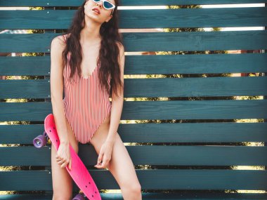 Güneş gözlüklü, genç, seksi, gülümseyen hippi kadın. Yazın mayo giyen moda kız. Pembe peni kaykayıyla çıldıran pozitif bir kadın. Çitin yanında poz veren bir model.