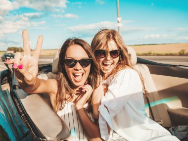 Üstü açık arabadaki iki güzel ve gülümseyen hippi kızın portresi. Cabriolet kullanan kaygısız seksi kadınlar. Pozitif modeller güneş gözlükleriyle geziyor ve eğleniyorlar.